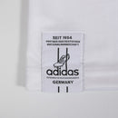 Vista frontal da camisa de futebol masculina branca da seleção da Alemanha, modelo torcedor home 2026-2027, com brasão bordado e detalhes da bandeira.