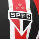Camisa oficial reserva (Away) do São Paulo FC 2025/26, versão Torcedor, da New Balance. A peça exibe as icônicas listras verticais vermelhas, pretas e brancas com cores profundas e nítidas. Apresenta gola redonda preta com detalhes em dourado metálico, que também decoram as bordas das mangas. O escudo do clube é aplicado em patch bordado de alta definição no lado esquerdo, contrastando com o logo da marca em dourado à direita. O tecido possui uma trama respirável e toque macio, ideal para o uso casual de al