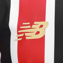 Camisa oficial reserva (Away) do São Paulo FC 2025/26, versão Torcedor, da New Balance. A peça exibe as icônicas listras verticais vermelhas, pretas e brancas com cores profundas e nítidas. Apresenta gola redonda preta com detalhes em dourado metálico, que também decoram as bordas das mangas. O escudo do clube é aplicado em patch bordado de alta definição no lado esquerdo, contrastando com o logo da marca em dourado à direita. O tecido possui uma trama respirável e toque macio, ideal para o uso casual de al