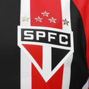 Camisa oficial de jogo (versão Jogador) do São Paulo FC 2025/26 New Balance. A peça exibe as tradicionais listras verticais em vermelho e preto, separadas por finas linhas brancas, com cores vibrantes e acabamento fosco. O escudo do clube e o logo da marca são aplicados em relevo emborrachado (heat-transfer) para maior leveza. A modelagem é visivelmente ajustada (slim fit), com gola em "V" adornada por detalhes dourados sutis, transmitindo uma estética de alta performance e sofisticação técnica.