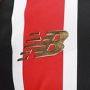 Camisa oficial de jogo (versão Jogador) do São Paulo FC 2025/26 New Balance. A peça exibe as tradicionais listras verticais em vermelho e preto, separadas por finas linhas brancas, com cores vibrantes e acabamento fosco. O escudo do clube e o logo da marca são aplicados em relevo emborrachado (heat-transfer) para maior leveza. A modelagem é visivelmente ajustada (slim fit), com gola em "V" adornada por detalhes dourados sutis, transmitindo uma estética de alta performance e sofisticação técnica.
