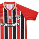 Camisa oficial de jogo (versão Jogador) do São Paulo FC 2025/26 New Balance. A peça exibe as tradicionais listras verticais em vermelho e preto, separadas por finas linhas brancas, com cores vibrantes e acabamento fosco. O escudo do clube e o logo da marca são aplicados em relevo emborrachado (heat-transfer) para maior leveza. A modelagem é visivelmente ajustada (slim fit), com gola em "V" adornada por detalhes dourados sutis, transmitindo uma estética de alta performance e sofisticação técnica.